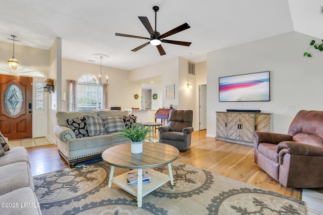 living room featuring vaulted ceiling, light wood finished floors, ceiling fan with notable chandelier, and visible vents