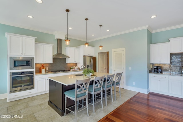 kitchen with stainless steel appliances, a kitchen bar, light countertops, a kitchen island with sink, and wall chimney exhaust hood