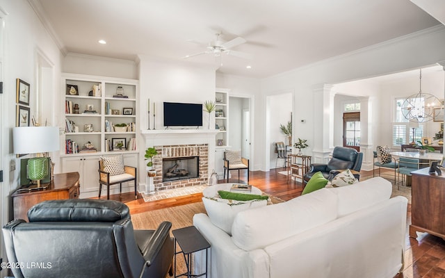 living room with ceiling fan with notable chandelier, ornamental molding, a brick fireplace, dark wood-type flooring, and built in shelves