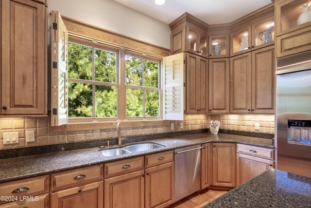 kitchen with dark stone counters, glass insert cabinets, appliances with stainless steel finishes, a sink, and backsplash