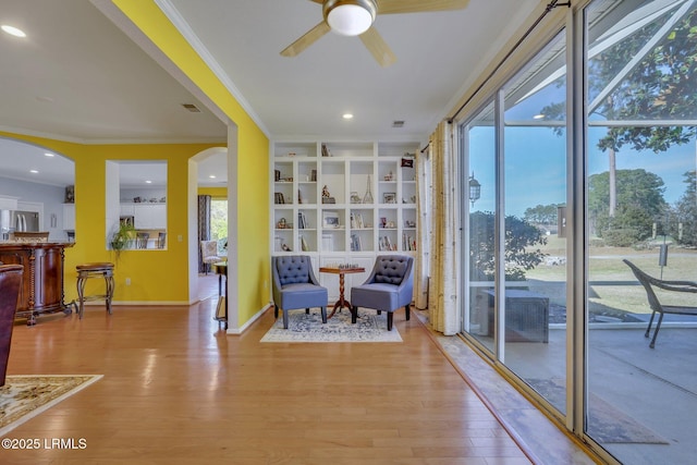 sitting room featuring built in shelves, ornamental molding, ceiling fan, and light wood-type flooring