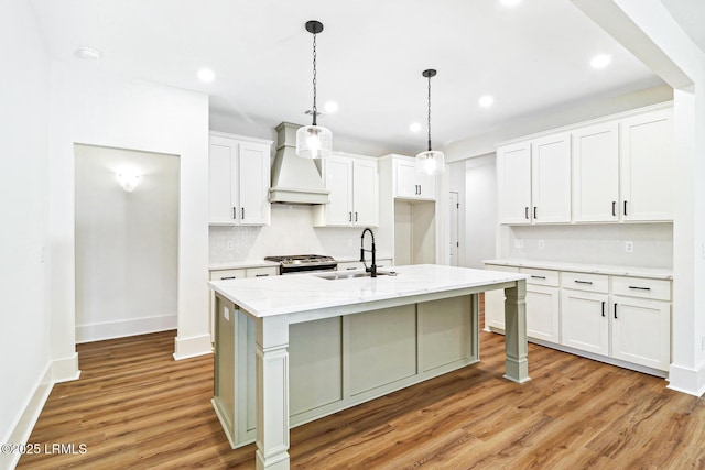 kitchen featuring premium range hood, sink, white cabinetry, hanging light fixtures, and a center island with sink