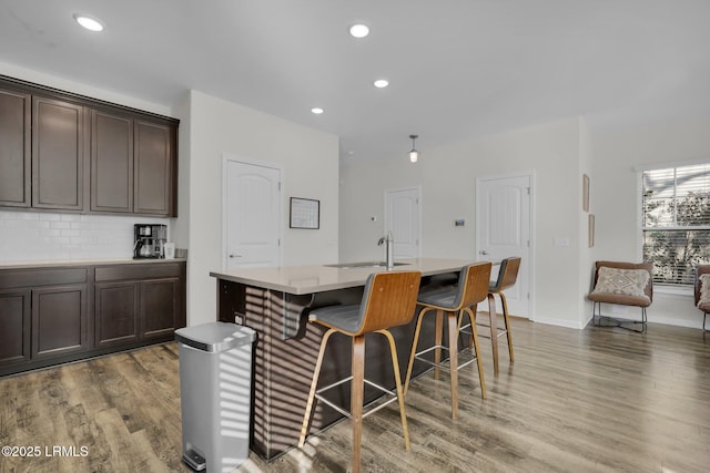 kitchen with backsplash, hardwood / wood-style floors, a kitchen breakfast bar, dark brown cabinetry, and an island with sink