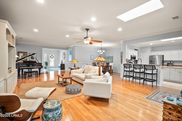 living room featuring crown molding, ceiling fan with notable chandelier, light wood-type flooring, and a skylight