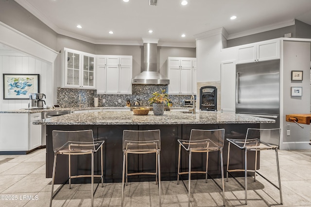 kitchen featuring a large island, decorative backsplash, stainless steel built in fridge, light stone countertops, and wall chimney exhaust hood