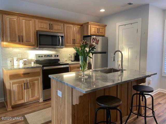 kitchen with sink, dark wood-type flooring, stainless steel appliances, light stone counters, and an island with sink