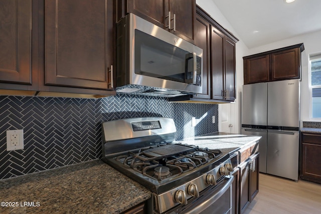 kitchen with dark stone countertops, vaulted ceiling, dark brown cabinetry, appliances with stainless steel finishes, and backsplash