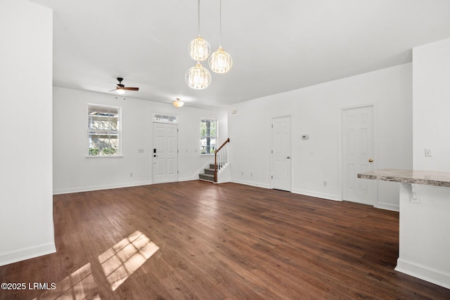 unfurnished living room with dark wood finished floors, ceiling fan with notable chandelier, stairs, and baseboards