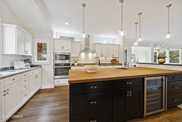 kitchen featuring wine cooler, wall chimney exhaust hood, butcher block counters, an island with sink, and stainless steel appliances