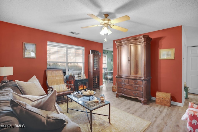 living room with ceiling fan, a wood stove, a textured ceiling, and light hardwood / wood-style flooring