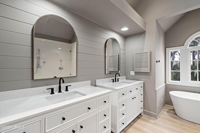 bathroom featuring wooden walls, wood finished floors, two vanities, a freestanding bath, and a tile shower