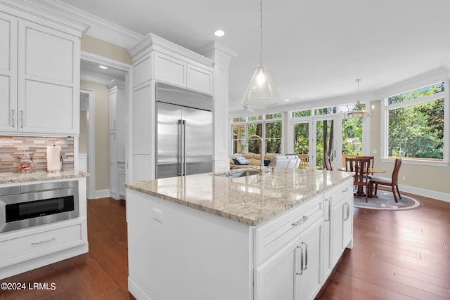 kitchen featuring decorative light fixtures, an island with sink, sink, white cabinets, and built in appliances