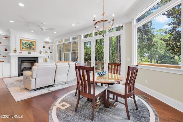 dining area featuring dark wood-type flooring, crown molding, a large fireplace, built in features, and ceiling fan with notable chandelier