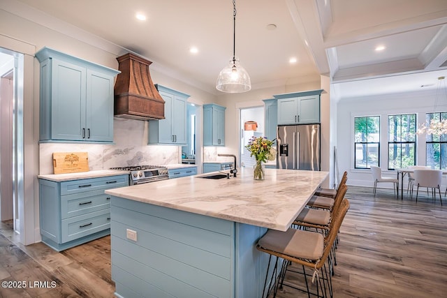 kitchen featuring sink, appliances with stainless steel finishes, hanging light fixtures, custom range hood, and a center island with sink