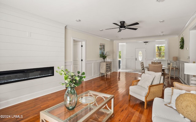 living area featuring a wainscoted wall, crown molding, a ceiling fan, a glass covered fireplace, and wood finished floors