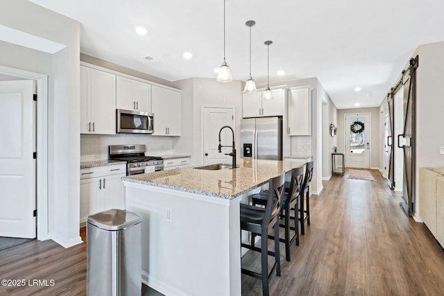 kitchen with sink, appliances with stainless steel finishes, white cabinets, a center island with sink, and a barn door