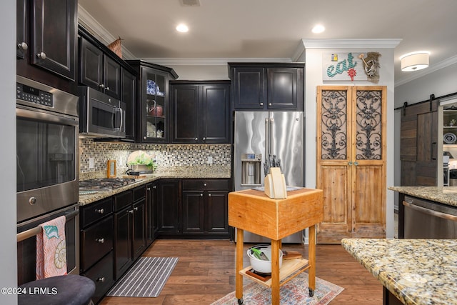 kitchen with dark wood-type flooring, light stone counters, ornamental molding, stainless steel appliances, and a barn door