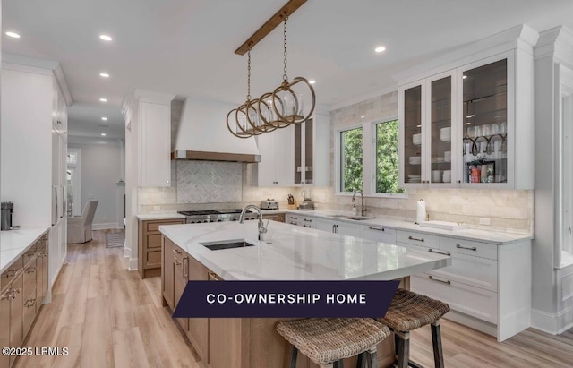 kitchen with sink, custom exhaust hood, white cabinetry, a center island with sink, and light stone countertops