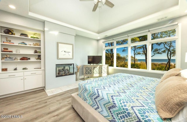 bedroom featuring ceiling fan, a multi sided fireplace, a tray ceiling, and light hardwood / wood-style floors