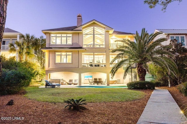 back house at dusk with a balcony, a lawn, and a patio area