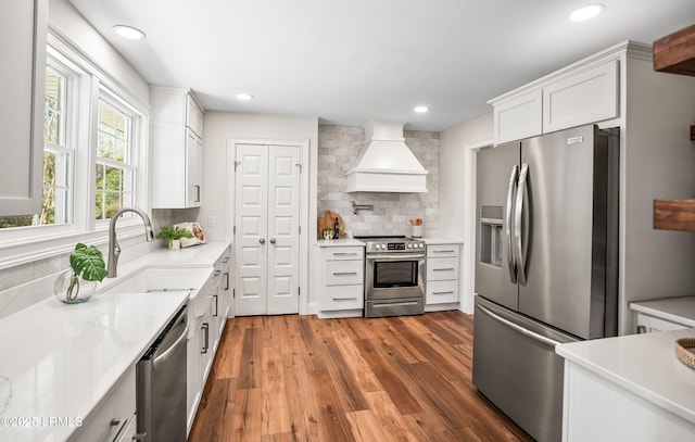 kitchen with decorative backsplash, white cabinets, custom range hood, appliances with stainless steel finishes, and a sink