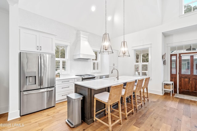 kitchen with sink, a kitchen island with sink, stainless steel appliances, custom range hood, and white cabinets