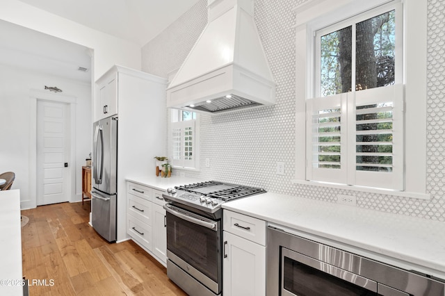 kitchen with white cabinetry, stainless steel appliances, custom range hood, decorative backsplash, and light wood-type flooring