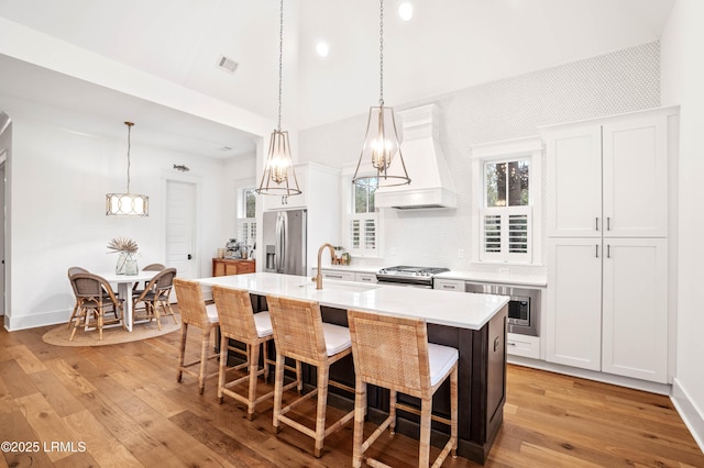 kitchen featuring pendant lighting, premium range hood, appliances with stainless steel finishes, white cabinets, and a center island with sink