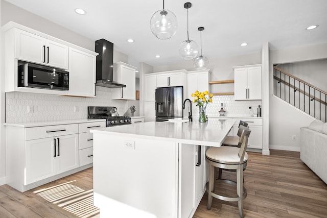 kitchen with appliances with stainless steel finishes, wall chimney exhaust hood, a kitchen island with sink, and light hardwood / wood-style flooring