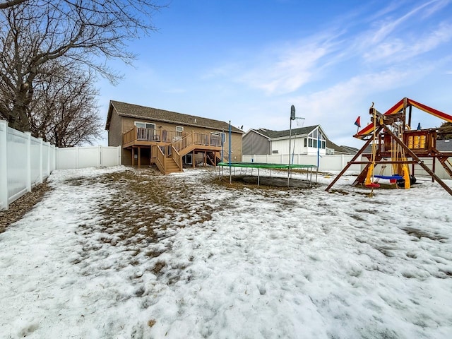 snow covered back of property featuring a deck, a playground, a fenced backyard, stairs, and a trampoline