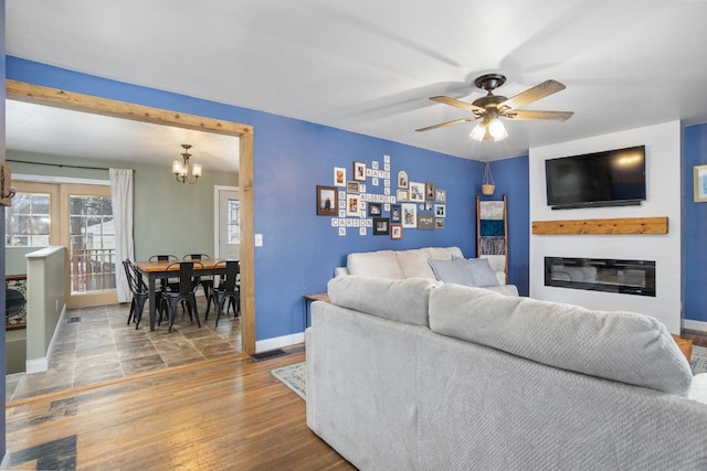 living room featuring visible vents, a glass covered fireplace, wood finished floors, baseboards, and ceiling fan with notable chandelier