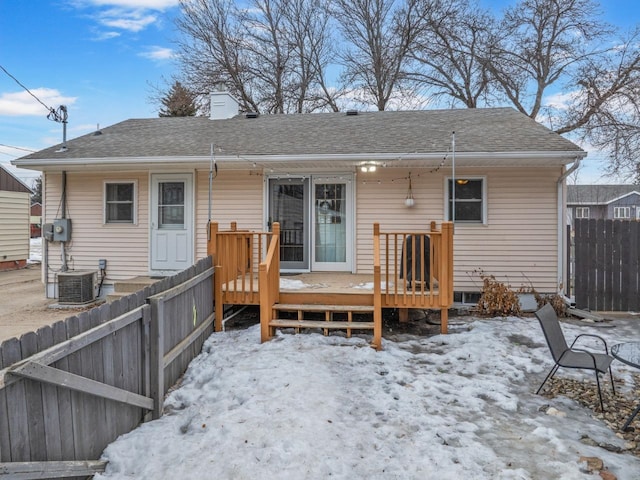 snow covered rear of property featuring a deck, central AC unit, a shingled roof, fence, and a chimney