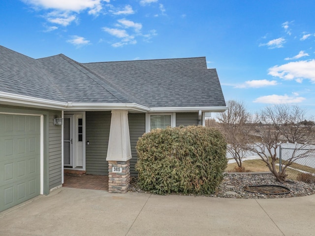 doorway to property featuring concrete driveway, roof with shingles, an attached garage, and fence