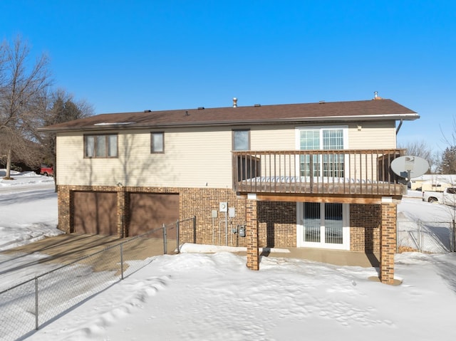 snow covered rear of property with driveway, a garage, a balcony, fence, and brick siding