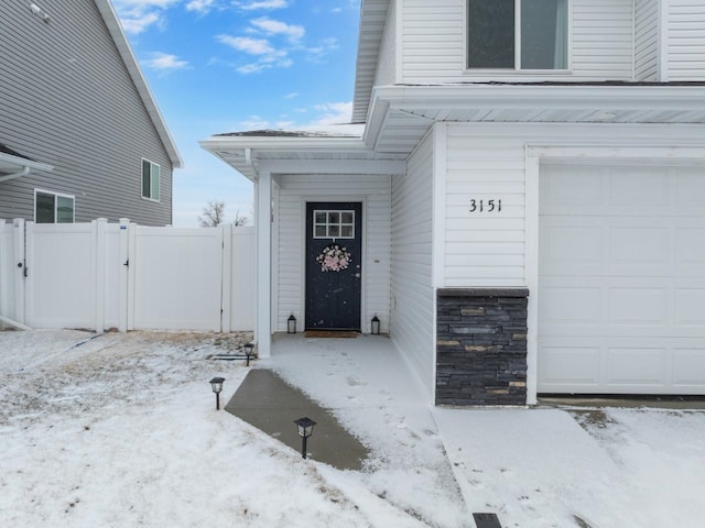 snow covered property entrance with a garage, stone siding, fence, and a gate