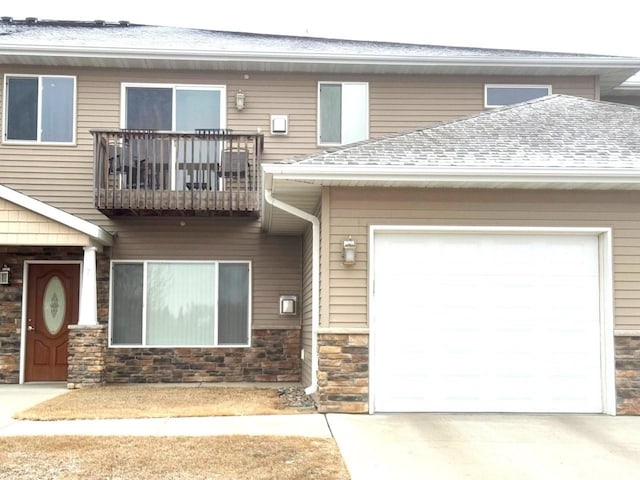 multi unit property with an attached garage, roof with shingles, a balcony, stone siding, and driveway