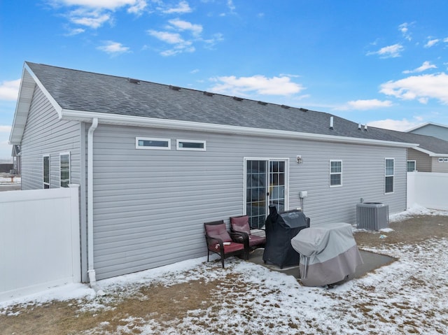 snow covered rear of property featuring central AC, fence, and roof with shingles