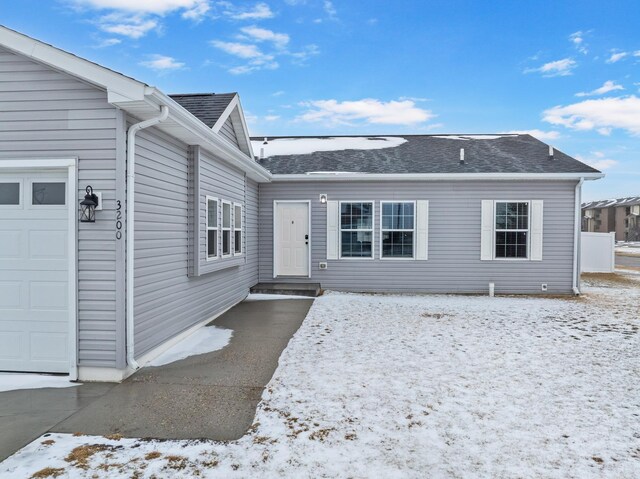 snow covered property entrance with an attached garage and roof with shingles