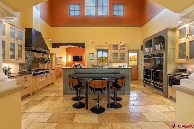 kitchen with a towering ceiling, wall chimney exhaust hood, backsplash, stainless steel gas stovetop, and a kitchen breakfast bar