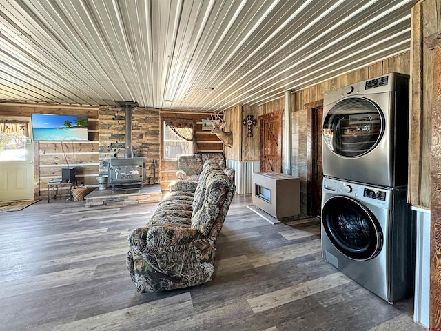 laundry room featuring dark hardwood / wood-style flooring, a wood stove, stacked washer / dryer, a healthy amount of sunlight, and wooden walls