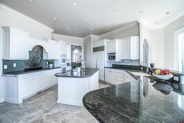 kitchen featuring dark stone counters, sink, crown molding, a center island with sink, and white cabinets