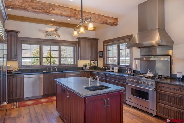 kitchen with beam ceiling, sink, stainless steel appliances, wall chimney range hood, and a center island with sink