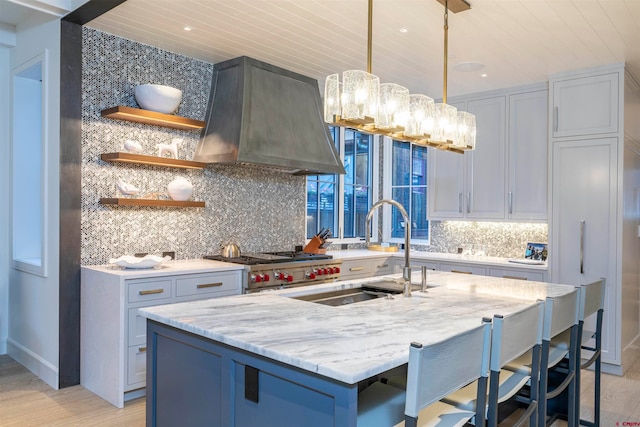 kitchen with wall chimney range hood, light hardwood / wood-style floors, hanging light fixtures, a center island with sink, and backsplash