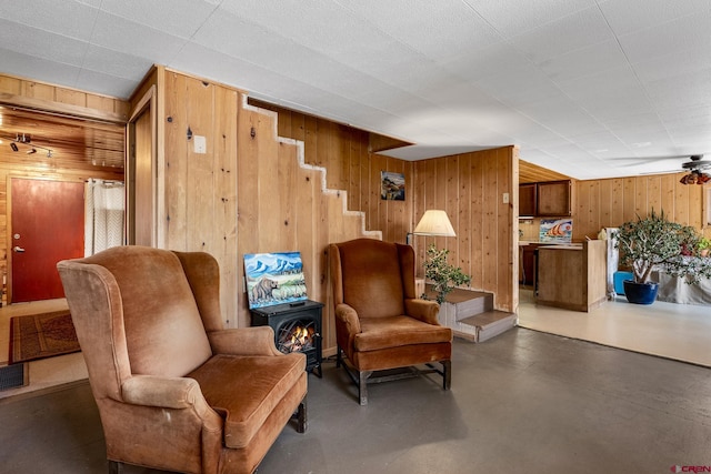 sitting room featuring a wood stove, wooden walls, ceiling fan, and concrete flooring