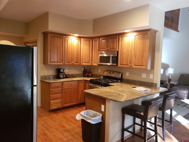 kitchen with kitchen peninsula, appliances with stainless steel finishes, light stone countertops, a kitchen breakfast bar, and light hardwood / wood-style flooring