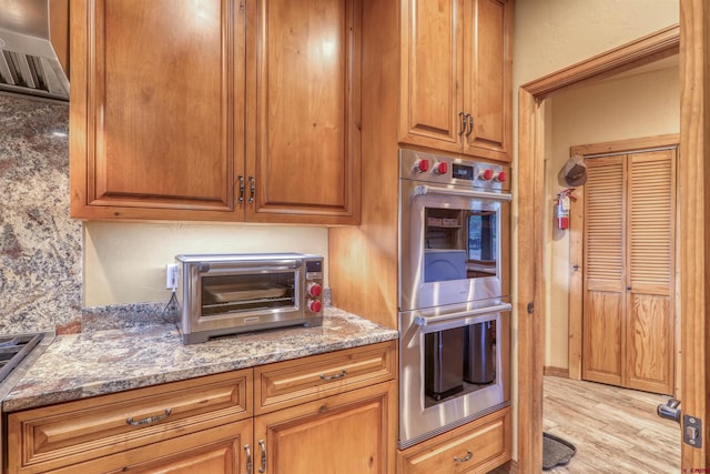 kitchen with double oven, light stone counters, exhaust hood, and light hardwood / wood-style floors