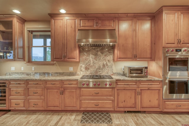 kitchen with exhaust hood, wine cooler, light hardwood / wood-style floors, light stone counters, and stainless steel appliances