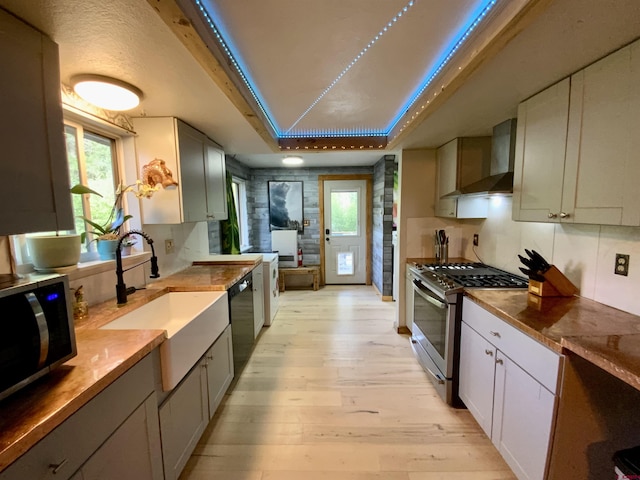 kitchen with stainless steel appliances, a sink, wall chimney range hood, light wood finished floors, and a raised ceiling