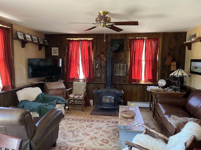 living room with wood walls, a wood stove, a wealth of natural light, ceiling fan, and a baseboard heating unit