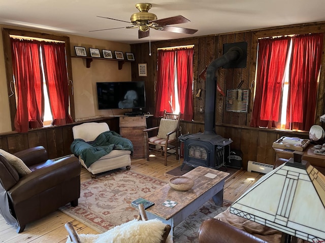 living room with wood walls, light hardwood / wood-style flooring, a wood stove, baseboard heating, and ceiling fan